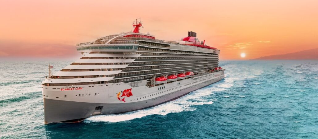 A large cruise ship from Virgin Voyages sails through the ocean under a colorful sunset sky. The ship's exterior, boasting significant upgrades, features multiple decks and rows of balconies, with lifeboats visible along the side. The water is calm, and the sun is setting on the horizon.