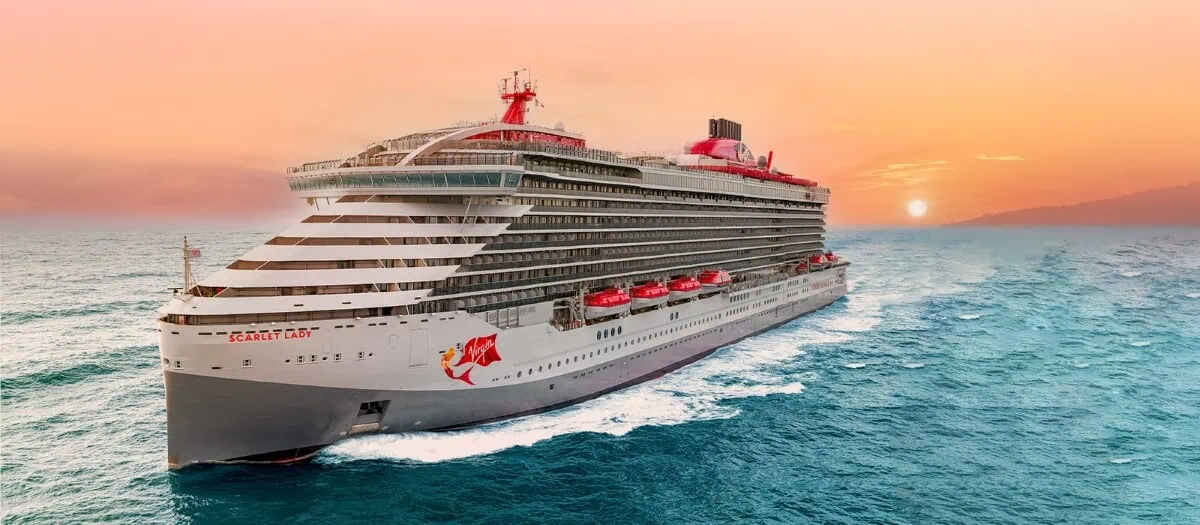 A large cruise ship from Virgin Voyages sails through the ocean under a colorful sunset sky. The ship's exterior, boasting significant upgrades, features multiple decks and rows of balconies, with lifeboats visible along the side. The water is calm, and the sun is setting on the horizon.