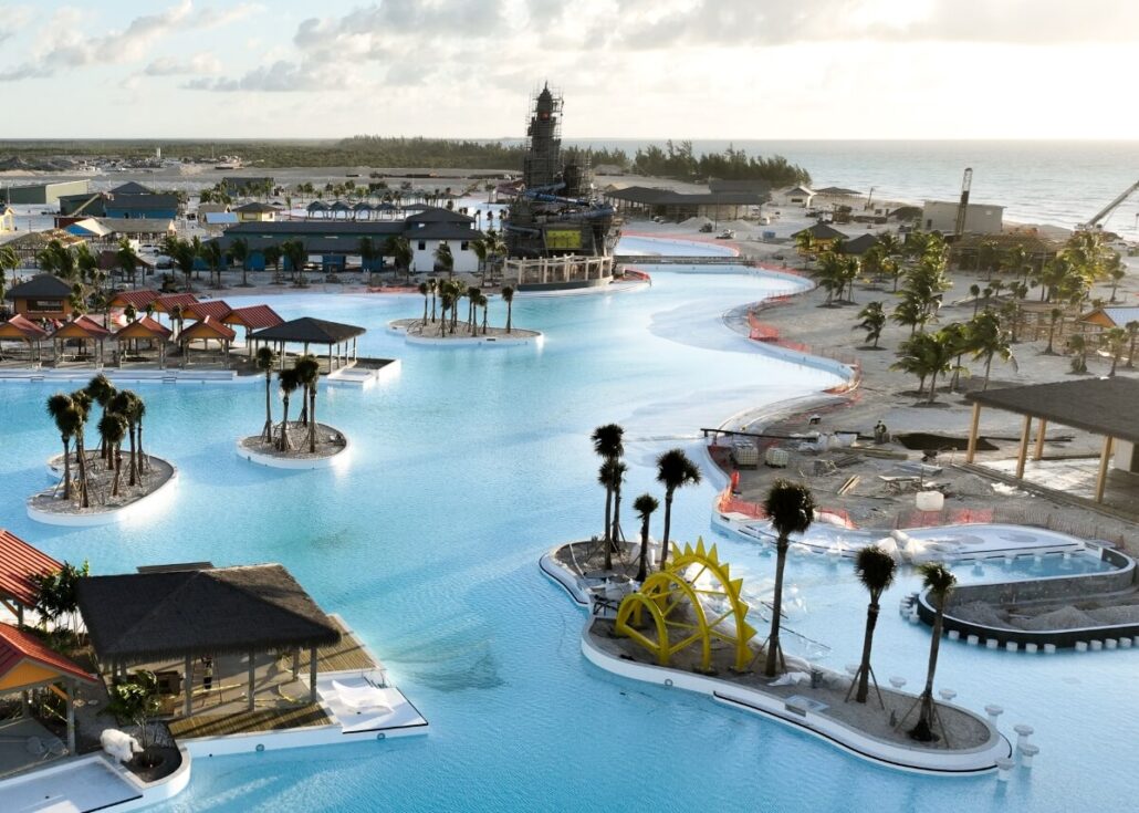 Aerial view of a large, winding pool with small islands and palm trees, reminiscent of a Carnival at The Bahamas. Shaded cabanas surround the pool, and a sun sculpture adorns one island. Celebration Key's construction is visible in the background, with the ocean shimmering in the distance.