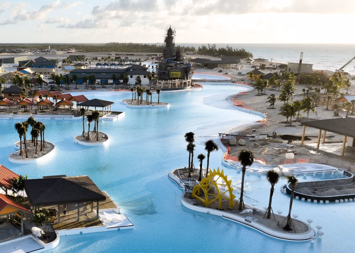 Aerial view of a large, winding pool with small islands and palm trees, reminiscent of a Carnival at The Bahamas. Shaded cabanas surround the pool, and a sun sculpture adorns one island. Celebration Key's construction is visible in the background, with the ocean shimmering in the distance.