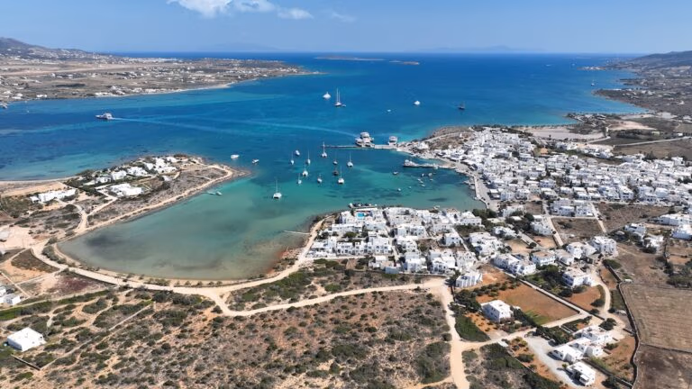 Aerial view of a coastal town with white buildings on the Greek island of Antiparos. The town is surrounded by turquoise water and a marina with sailboats, hinting at luxury cruises. The landscape blends greenery and dry terrain, with more land visible in the distance under a clear sky.