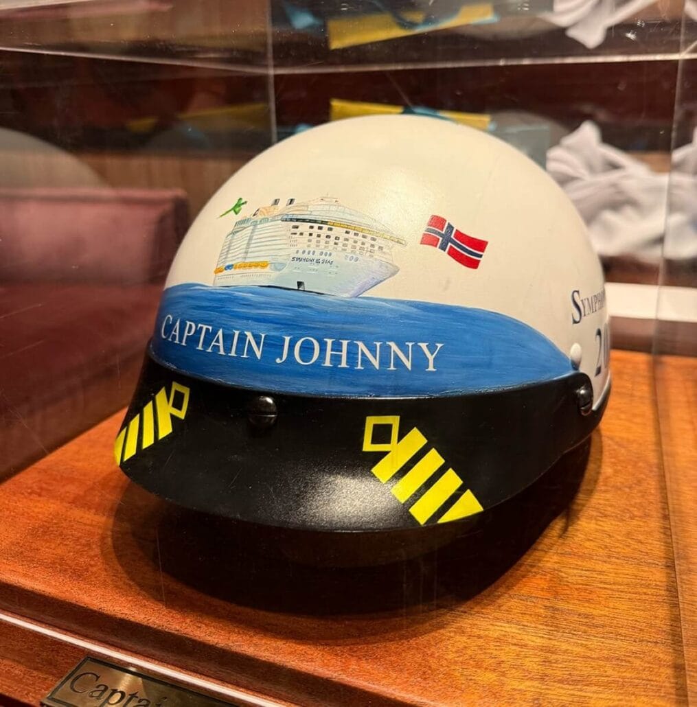 A white helmet showcases a painted cruise ship and a Norwegian flag, with "Captain Johnny Faevelen" inscribed on a blue band. Yellow striped patterns adorn the black section, capturing maritime elegance. Displayed in a glass case, this tribute resonates with the spirit of dedicated captaincy.