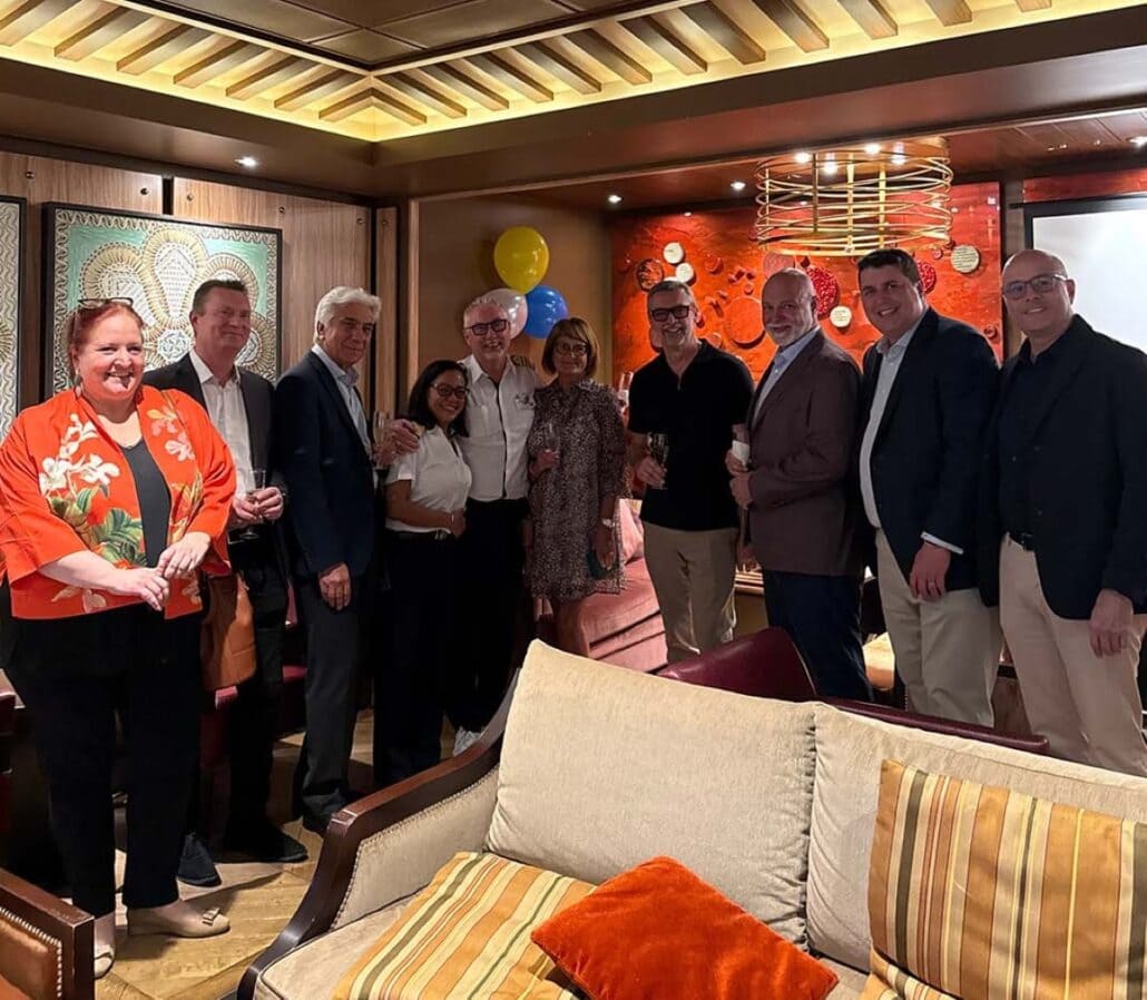 A group of nine people, including Johnny Faevelen, is posing indoors. They stand close together, some holding drinks. Behind them are colorful balloons and decorative wall art. The room has warm lighting and wooden accents. Everyone is dressed in semi-formal attire, like a captain's gala night.
