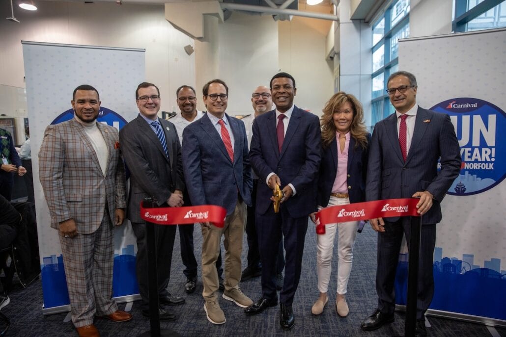 A group of eight people stands together indoors at a ceremonial event. Two individuals in the center hold a ribbon and a large pair of scissors, under banners that read "Carnival" and "Fun Year! Norfolk." It's a nod to Carnival Cruise Line's exciting year-round cruises launching from Norfolk.
