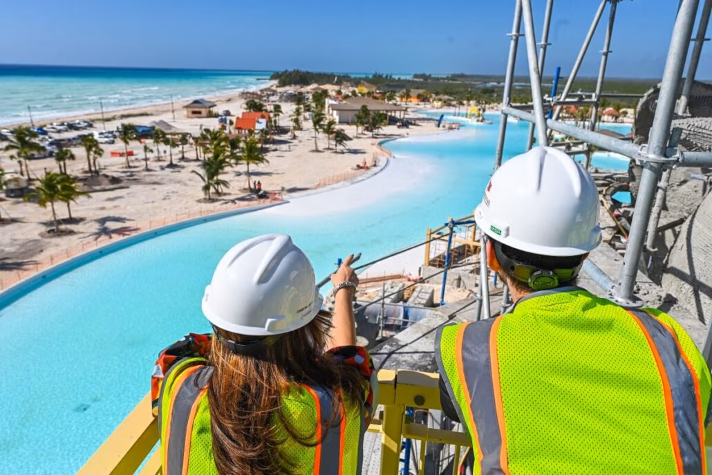 Two construction workers in safety vests and helmets stand on scaffolding overlooking a large pool project at a beach. One worker points toward the site, envisioning the next Carnival Celebration. The ocean, sandy beach, and palm trees offer a taste of the Bahamas in the background.