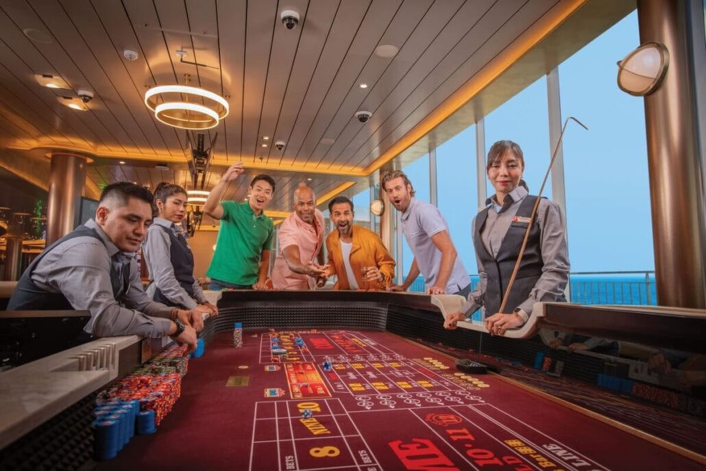 A group of people huddle around a casino table on a cruise ship, the room well-lit and lively. Chips are stacked high as they engage in offline gambling, with a dealer poised to the side. Large windows offer stunning ocean views beyond the cruise casinos' confines.