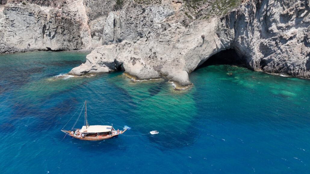 A small wooden sailboat with a canopy floats in the clear blue sea near rocky cliffs and a large cave opening, reminiscent of a serene cruise stop. The boat features a Greek flag and has a small dinghy attached, all set against the stunning backdrop of Antiparos's varying blue and green waters.