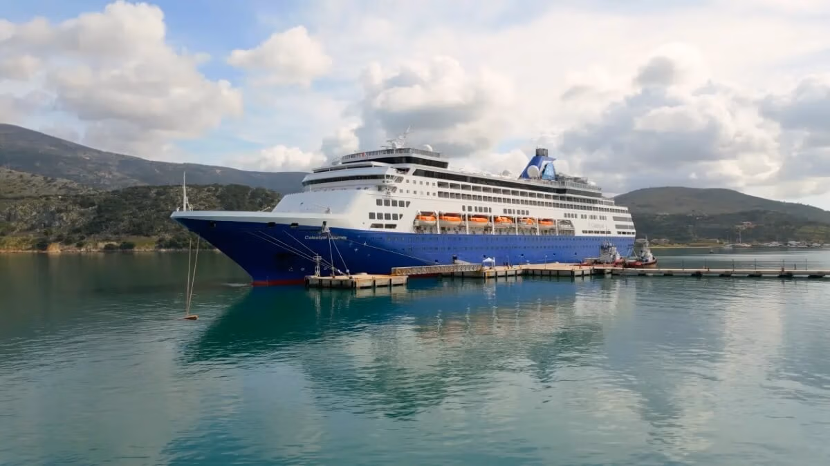A large blue and white cruise ship, the Celestyal Journey, is docked at a pier on calm green waters. The vessel, fresh from its $6.8 million refit, displays multiple decks with lifeboats hanging on the side. Rolling hills and partly cloudy skies provide a picturesque backdrop.