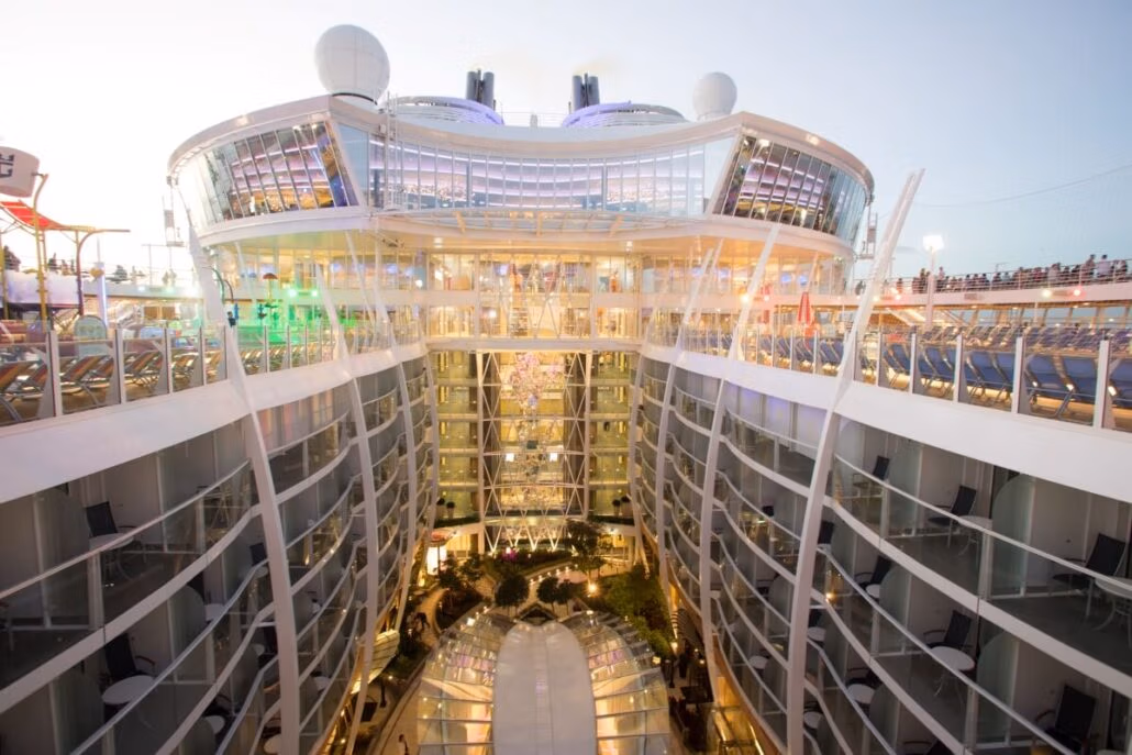 A glimpse inside a Royal Caribbean cruise ship reveals multiple balconies overlooking an open central area adorned with trees and lights. The modern design showcases glass and metal elements, with the sky peeking through in the background, ensuring both style and stellar CDC sanitation inspection standards.