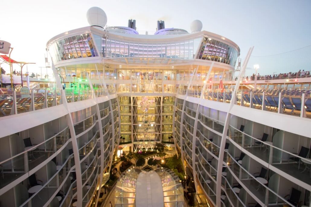 A glimpse inside a Royal Caribbean cruise ship reveals multiple balconies overlooking an open central area adorned with trees and lights. The modern design showcases glass and metal elements, with the sky peeking through in the background, ensuring both style and stellar CDC sanitation inspection standards.