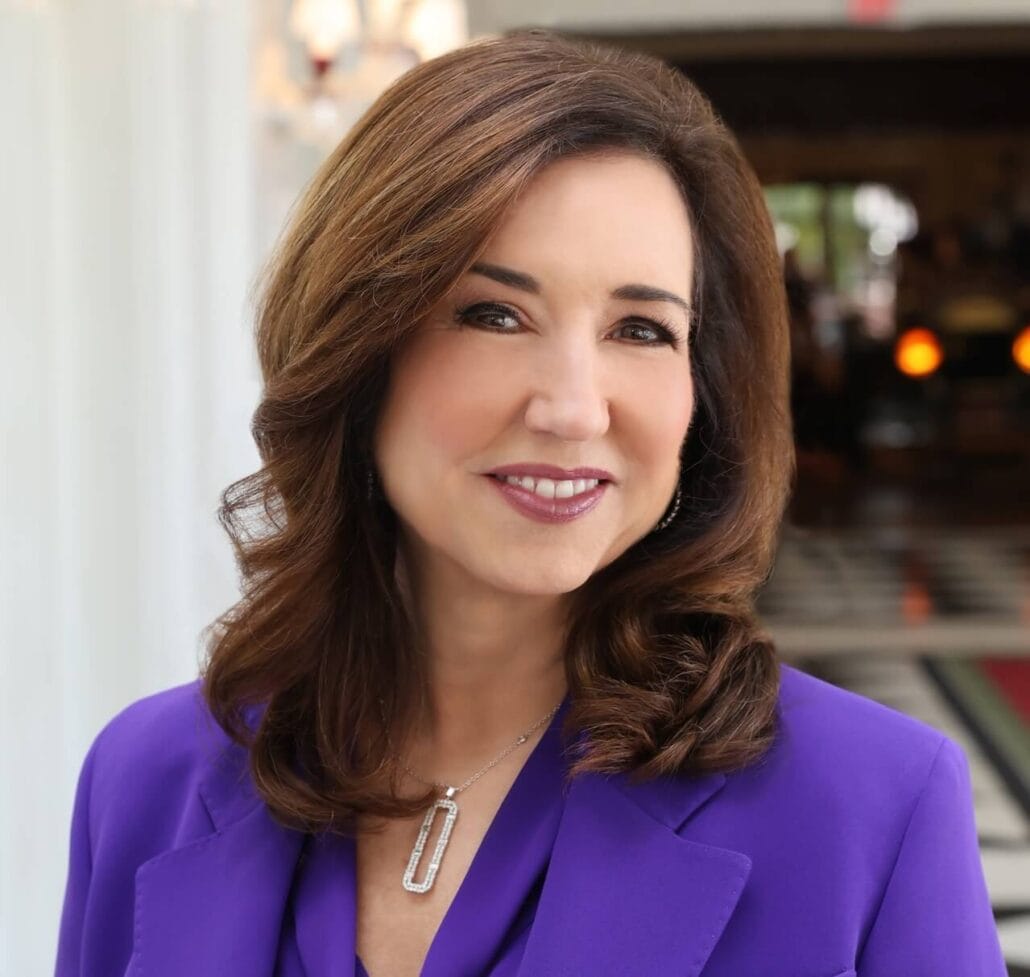 A woman with shoulder-length brown hair is smiling, evoking the warmth of a Carnival Cruise Line voyage. She is wearing a purple blazer and a necklace with a rectangular pendant. The softly blurred background highlights a black and white floor under cozy lighting.