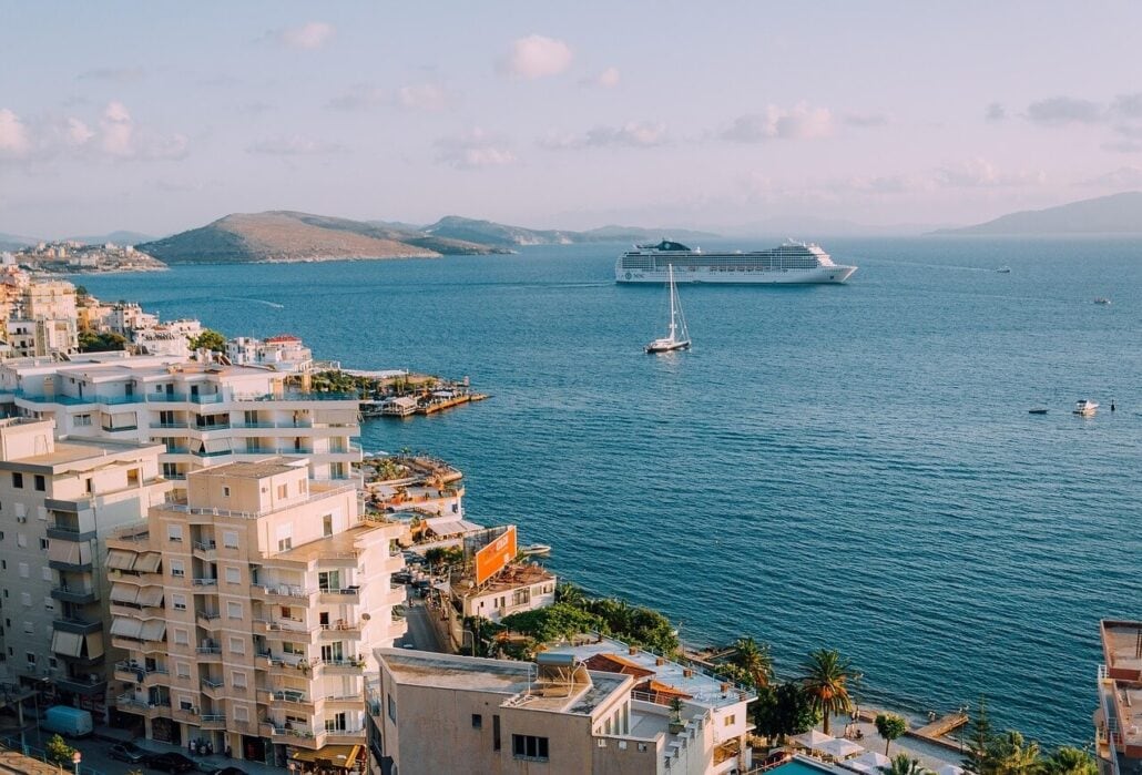 View of a coastal city with several white buildings in the foreground. A large cruise ship, reminiscent of a swindling scam artist's escapade, and a small sailboat grace the blue sea. Hills are visible in the background under a clear sky.