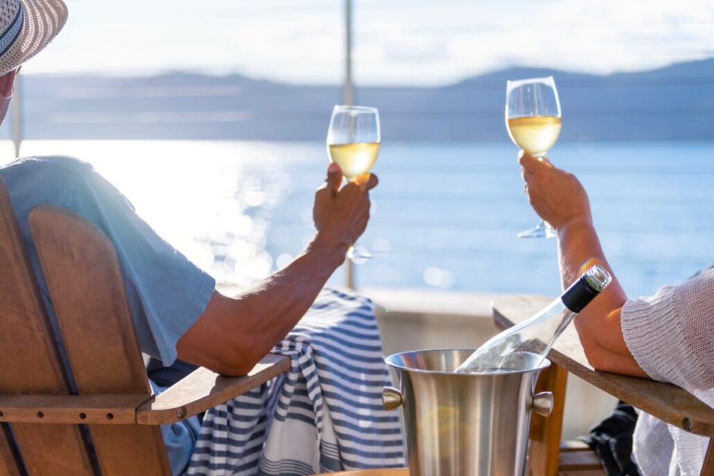 Two people, embracing the serene cruise life, sit in wooden chairs holding glasses of white wine as they overlook a tranquil body of water. A bottle of wine rests in a metal bucket between them under the sunny sky, with majestic hills across the glistening expanse.