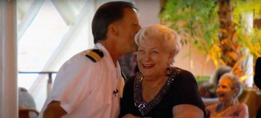 A person in a captain's uniform kisses an elderly retiree on the cheek. The woman is smiling and wearing a black dress with decorative trim, as if just back from cruise life. They are indoors with blurred people and plants in the background.