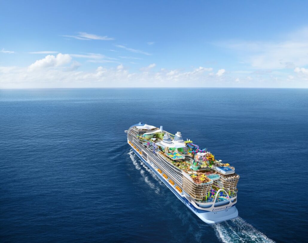 The Icon-Class ship from Royal Caribbean gracefully sails on a calm ocean under a blue sky with scattered clouds. It boasts multiple decks adorned with colorful attractions, including a water park and outdoor amenities, as it cruises forward on the open sea.