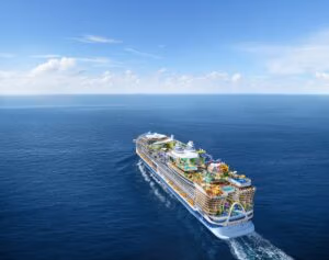 The Icon-Class ship from Royal Caribbean gracefully sails on a calm ocean under a blue sky with scattered clouds. It boasts multiple decks adorned with colorful attractions, including a water park and outdoor amenities, as it cruises forward on the open sea.