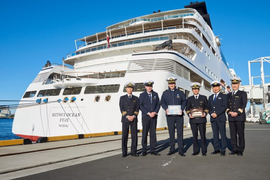 Six uniformed men stand proudly in front of the large white cruise ship, "Mitsu Ocean Fuji Nassau," docked at the port. This Japanese luxury cruise offers North Americans an unforgettable journey. The clear, blue sky hints at a perfect sunny day for setting sail.