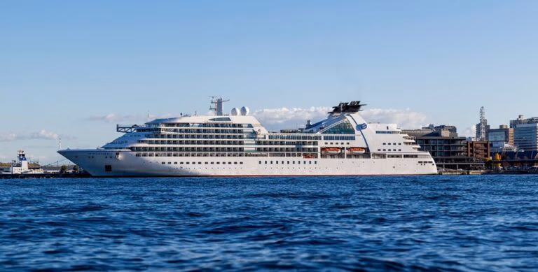 A large white cruise ship from a renowned Japanese luxury cruise line is docked at the pier, with multiple decks and lifeboats visible. Modern buildings and industrial structures rise in the background under a clear blue sky. The calm water reflects this scene of leisurely sailings for North Americans.