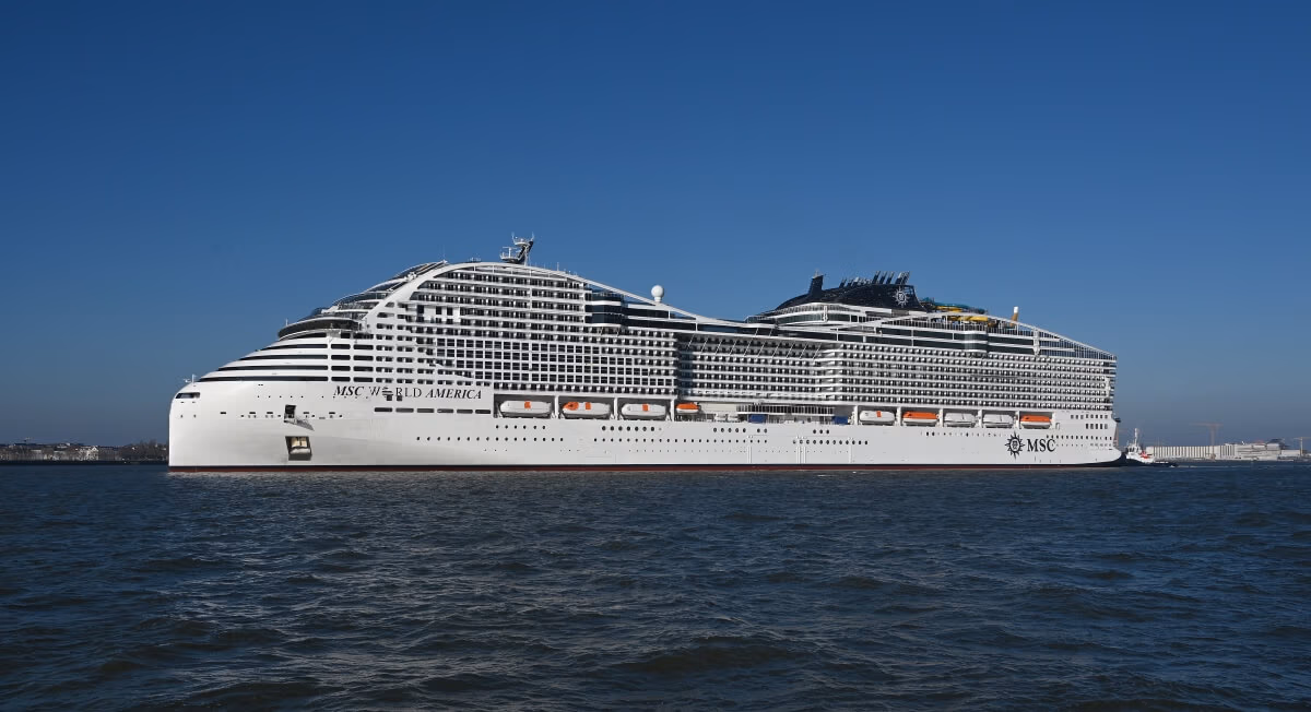 A large white cruise ship, reminiscent of the grandeur of MSC World America, glides gracefully across a calm body of water under a clear blue sky. With multiple decks lined with lifeboats and various top-deck structures, it appears ready for its upcoming U.S. debut after successful sea trials.
