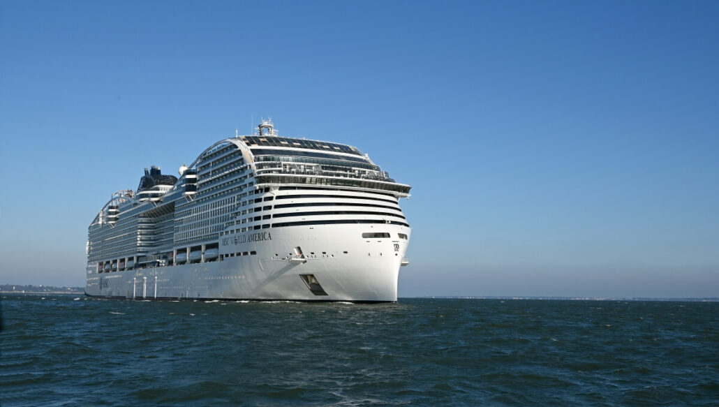 The MSC World America, making its U.S. debut, sails gracefully on a calm sea under a clear blue sky. This large white cruise ship with multiple decks is in the foreground, while distant land beckons on the left.