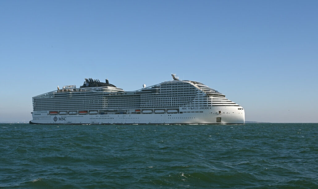 The MSC World America, making its U.S. debut, sails on a calm sea against a clear blue sky. The large white cruise ship boasts multiple decks and numerous windows, gliding from right to left with subtle waves around it.