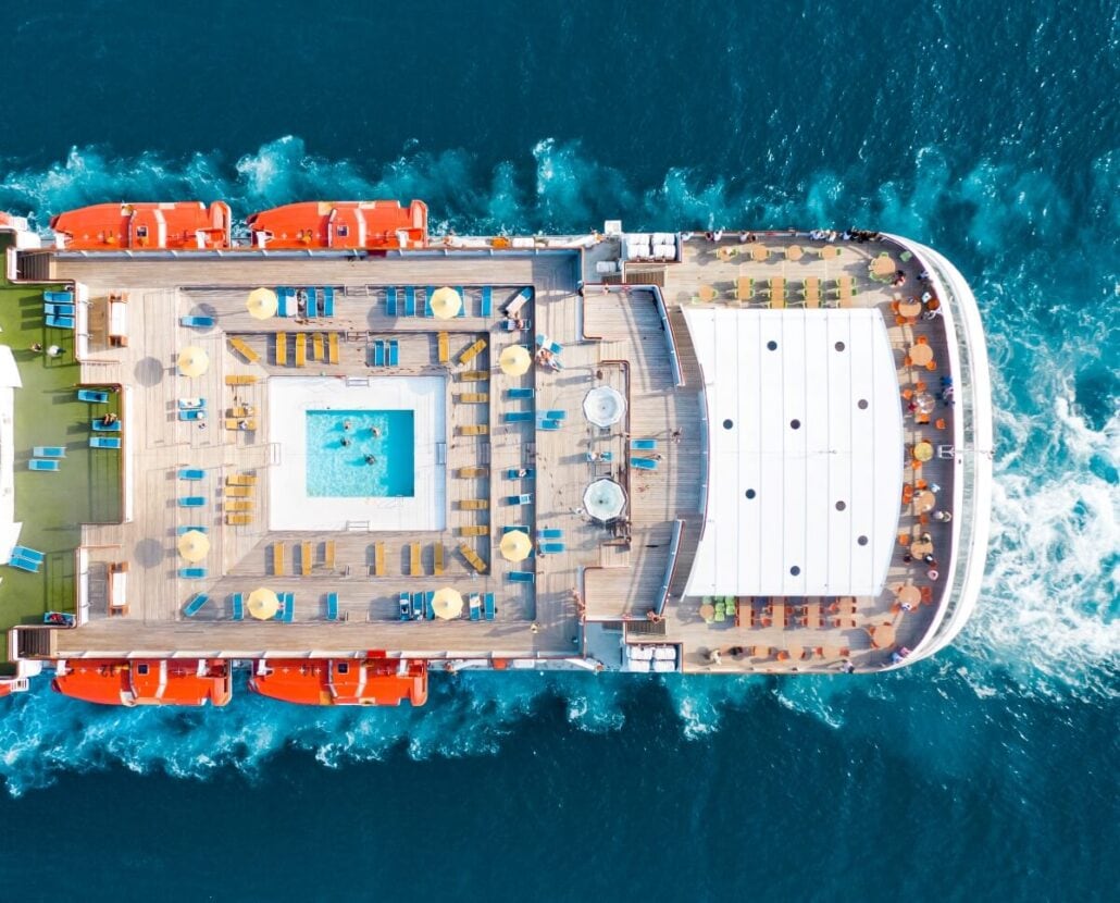 Aerial view of a cruise ship's deck in Sea Paradise, showcasing a rectangular swimming pool encircled by sun loungers and tables with umbrellas. The ship sails through blue water, leaving a white wake behind. Bright and sunny weather enhances this maritime escape.