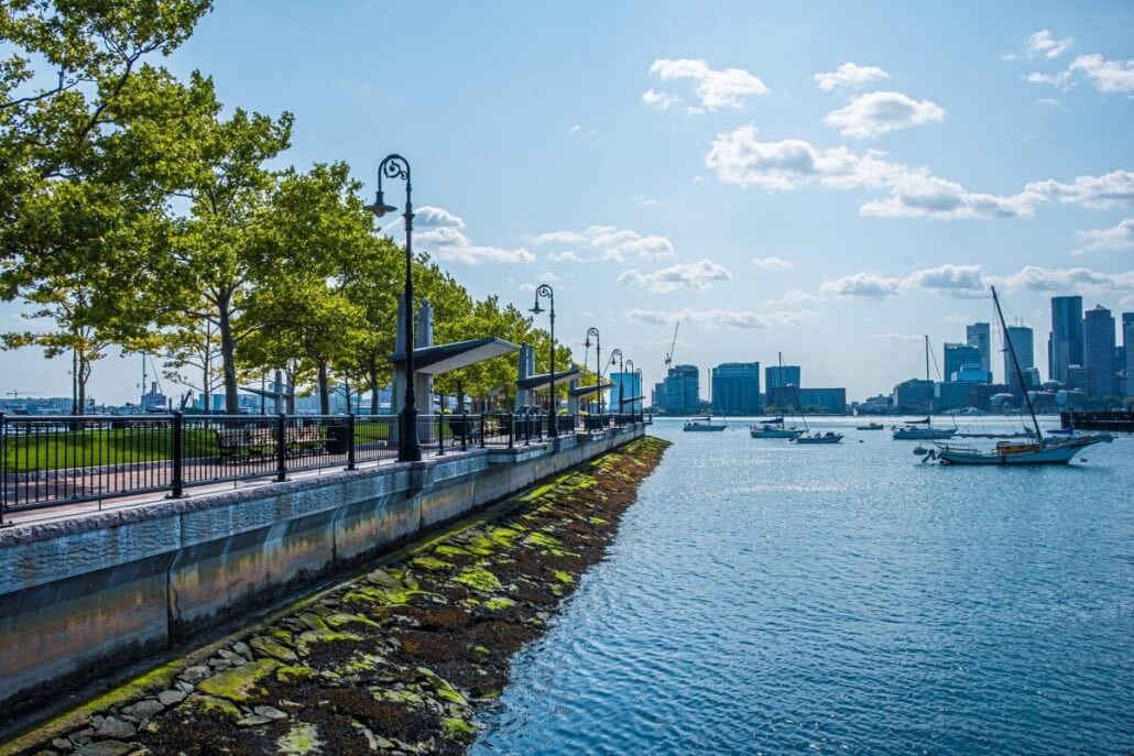 A waterfront scene in Boston 2026 features a concrete walkway lined with street lamps and trees on the left. Boats, soon embarking on their cruise, float in the water on the right, with a city skyline as the backdrop. The sky is clear with some clouds.
