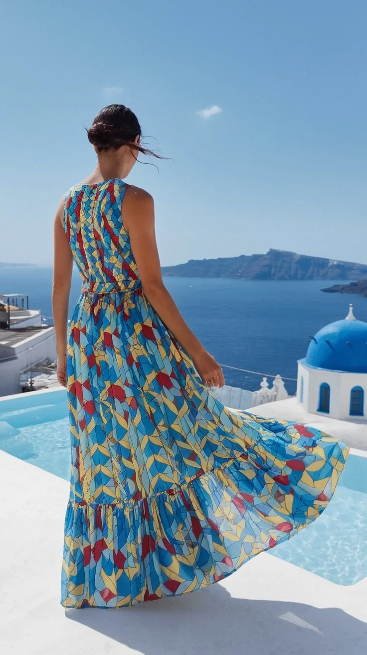 A person in a colorful geometric-patterned dress stands by a pool overlooking the blue sea and distant cliffs, embodying chic outfit ideas for a Mediterranean cruise. A white building with a blue dome is visible under the clear sky, as their hair is tied back and the dress flows with the breeze.