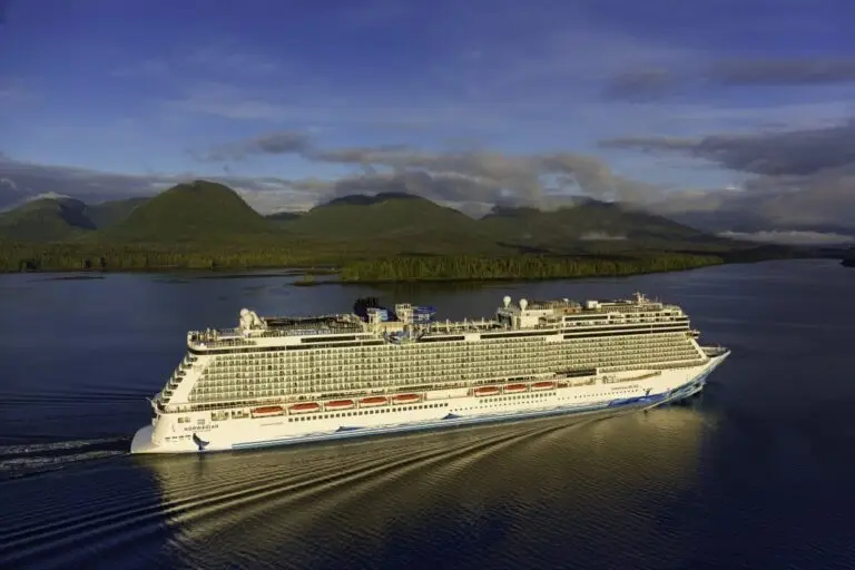 A large white cruise ship from Norwegian Cruise Line sails through calm waters, surrounded by a scenic landscape of green, forested mountains under a cloudy sky.