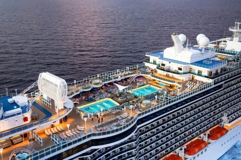 Aerial view of the Sky Princess cruise ship's deck at sunset. The deck features multiple swimming pools, numerous sun loungers, hot tubs, and life boats. The ship sails on calm waters, with warm lighting illuminating the recreational areas.