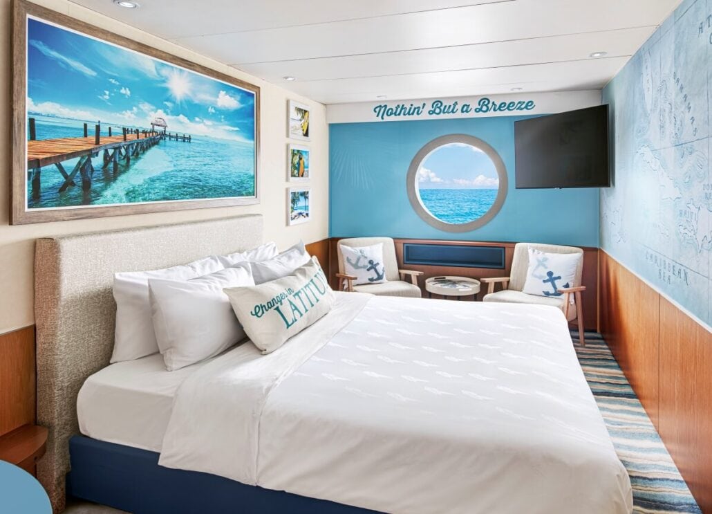 A cozy bedroom at Margaritaville at Sea features a large bed with white linens, under a photograph of a pier over turquoise water. There's a round window with an ocean view, two chairs, and a wall map. A pillow reads "Change in Latitude." The room has a nautical theme throughout.