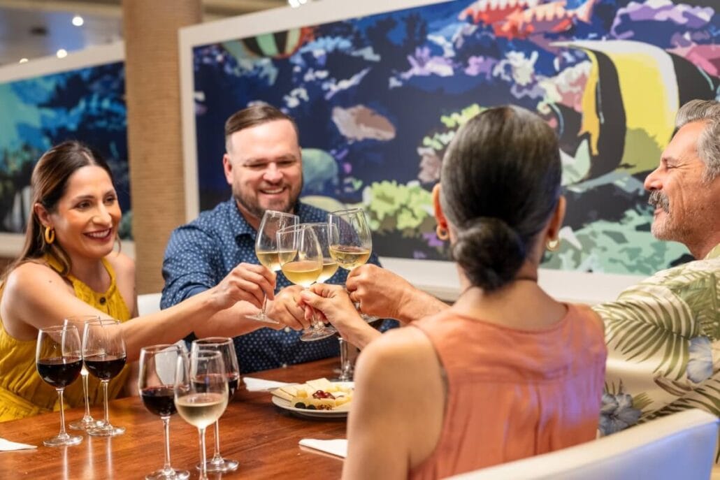 Four people clink wine glasses at a dining table, enjoying the vibrant ambiance of Margaritaville at Sea. Two women and two men are seated, smiling, against a backdrop of colorful marine art. As they chat over their red and white wines, the exceptional onboard service adds to their delightful evening.