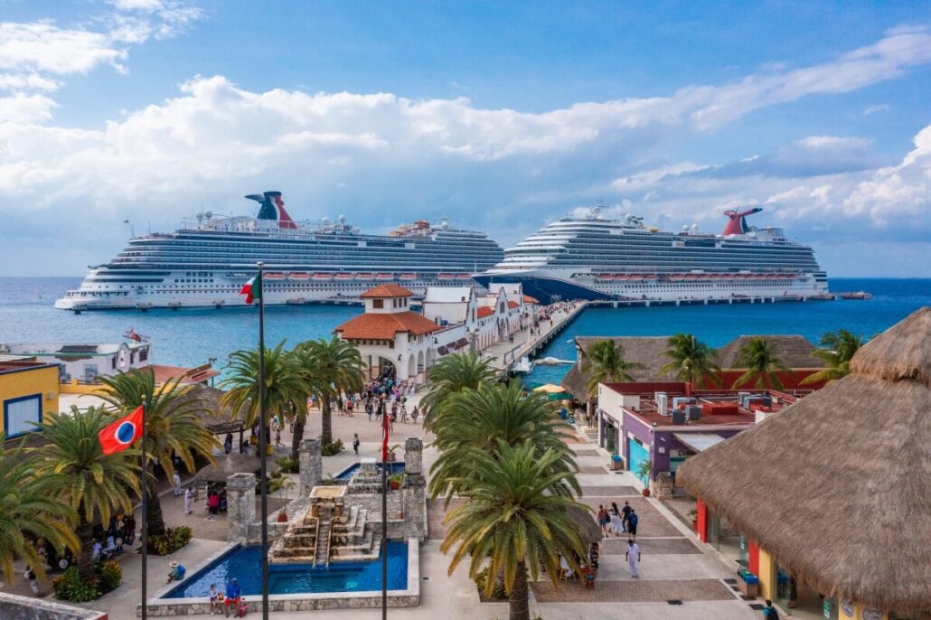 Experience a scenic view of the bustling cruise port, where two large Carnival Cruises ships are docked. Amidst palm trees and colorful buildings, a lively crowd gathers near the fountain under a partly cloudy sky, while the ocean's vibrant blue enchants every visitor.