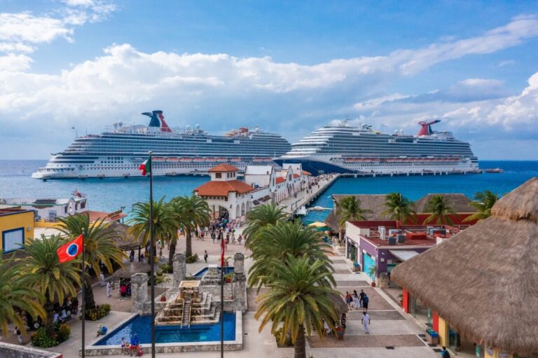 Experience a scenic view of the bustling cruise port, where two large Carnival Cruises ships are docked. Amidst palm trees and colorful buildings, a lively crowd gathers near the fountain under a partly cloudy sky, while the ocean's vibrant blue enchants every visitor.