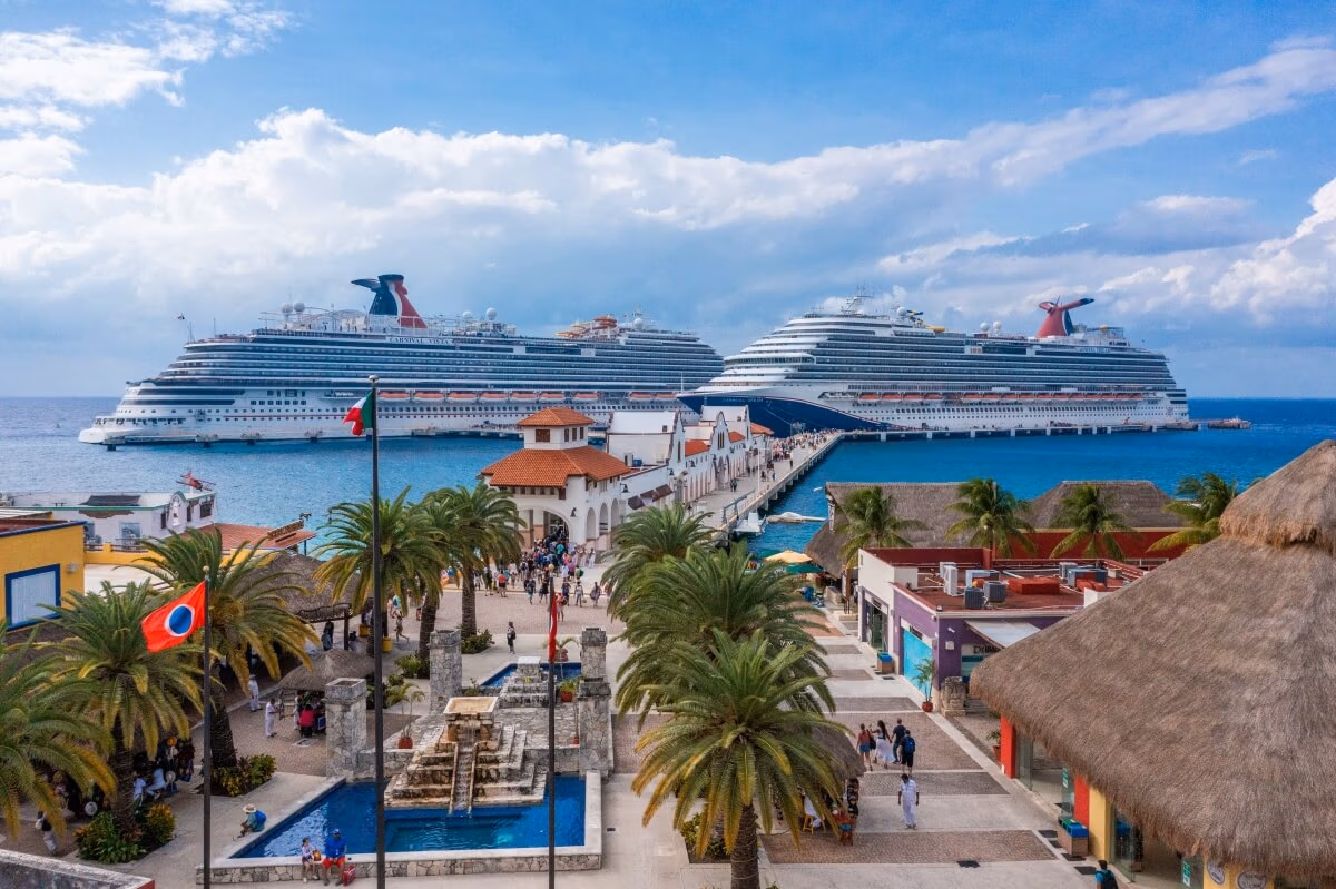 Experience a scenic view of the bustling cruise port, where two large Carnival Cruises ships are docked. Amidst palm trees and colorful buildings, a lively crowd gathers near the fountain under a partly cloudy sky, while the ocean's vibrant blue enchants every visitor.