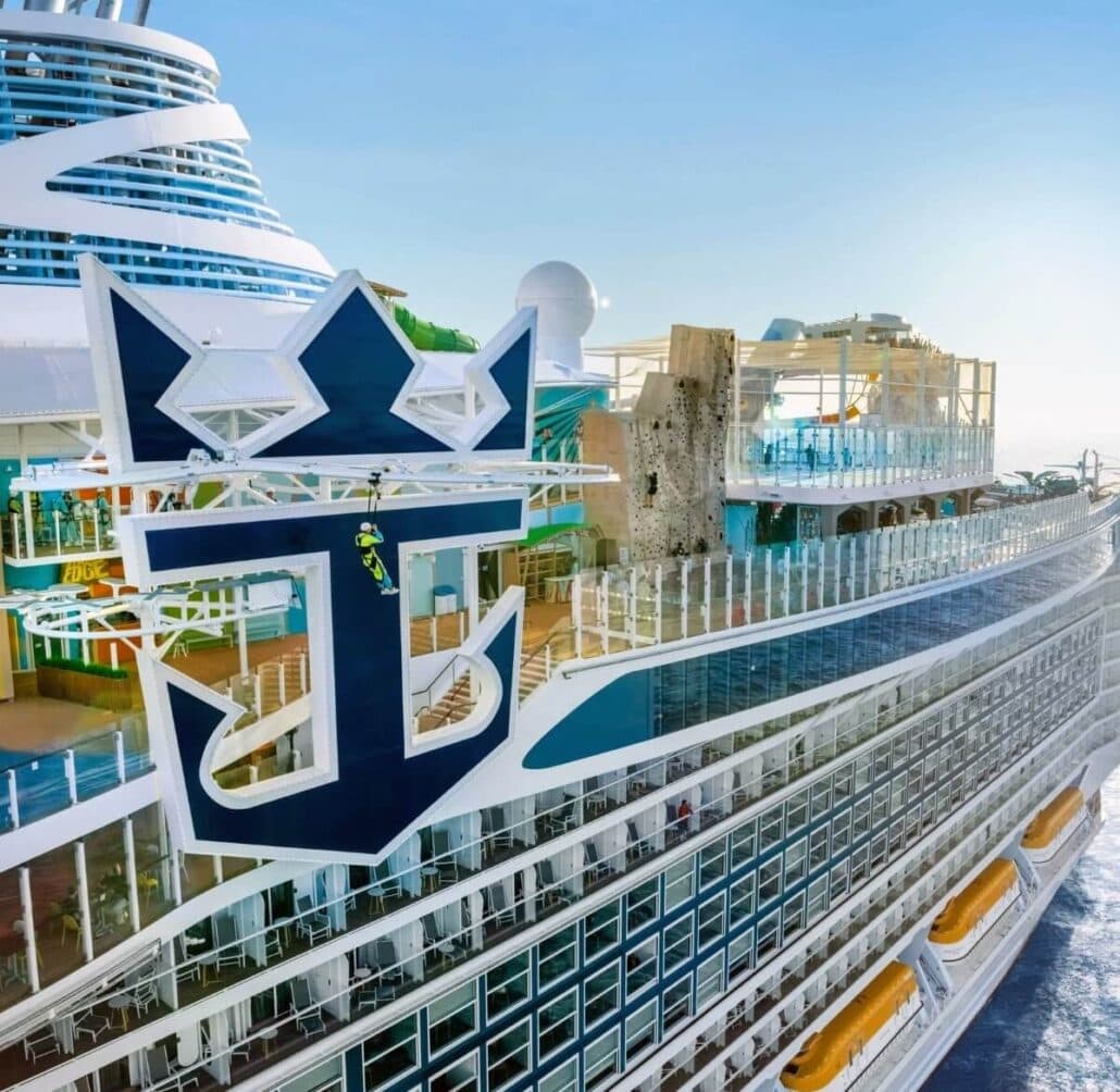 A Royal Caribbean cruise ship with a visible emblem graces the sea, boasting multiple decks, outdoor seating areas, and a rock climbing wall. The sky is clear as this majestic vessel docks or sails smoothly through serene waters.