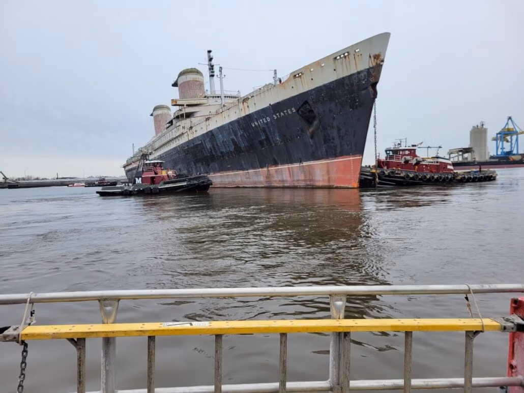 An aged, legendary ocean liner with visible rust and peeling paint is being carefully navigated by tugboats on its final voyage through the harbor. The ship’s name is faintly visible, while the foreground features a yellow and white metal railing.
