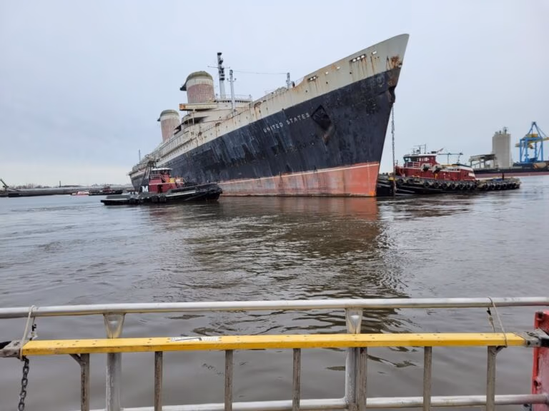 An aged, legendary ocean liner with visible rust and peeling paint is being carefully navigated by tugboats on its final voyage through the harbor. The ship’s name is faintly visible, while the foreground features a yellow and white metal railing.