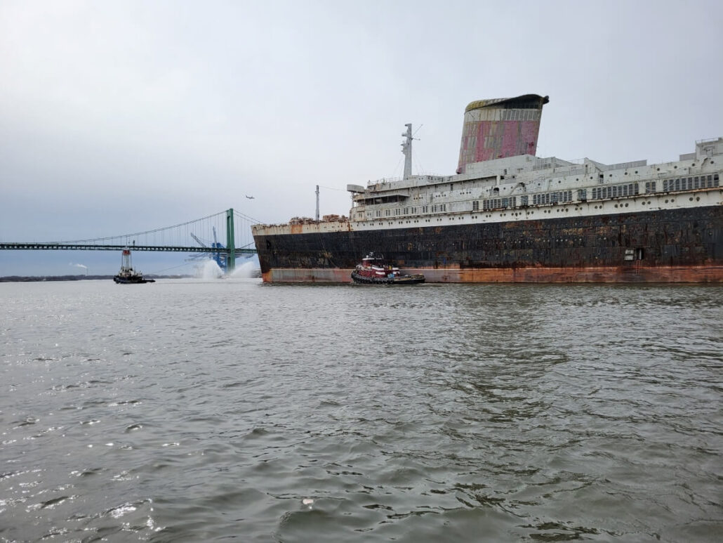 The legendary ocean liner, marked by rust and faded paint, drifts through the water as two tugboats maneuver nearby. On its final voyage, a jet of water sprays from the vessel under an overcast sky, with a bridge standing in the background.