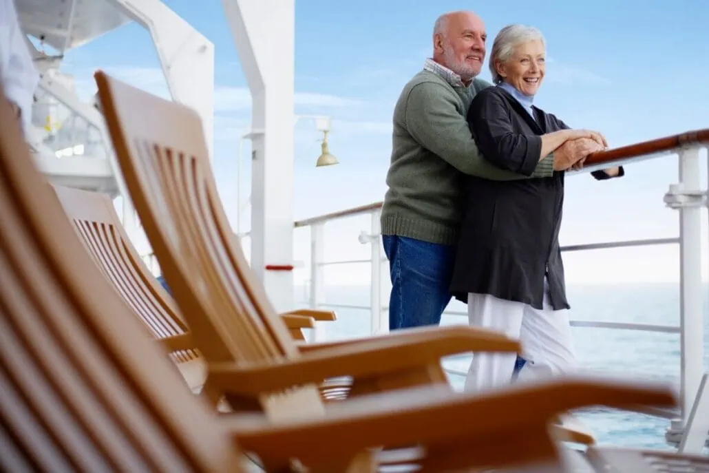 An older couple stands on a cruise ship deck, embodying the essence of Cruise Life. The man embraces the woman from behind as they lean on a railing, both smiling warmly at the vast ocean. Empty wooden deck chairs sit peacefully in the foreground, echoing a serene escape for retirees.