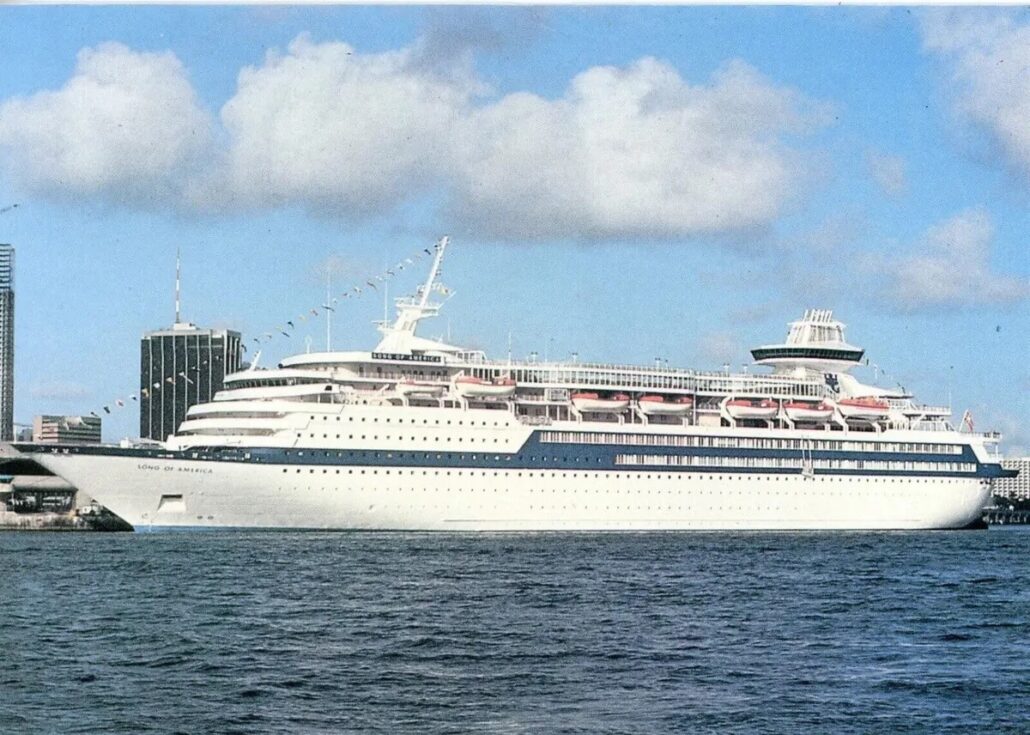 A large white cruise ship, the "Song of America," from Royal Caribbean, is docked at a port. This majestic vessel boasts multiple decks and visible lifeboats. In the background, a city skyline with tall buildings rises under a partly cloudy sky.