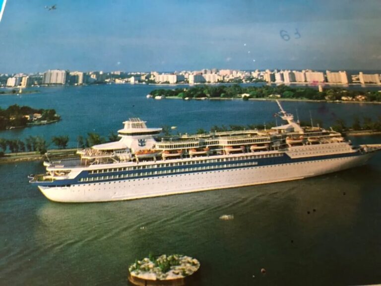 A large white Royal Caribbean cruise vessel gracefully sails through a calm body of water, with a cityscape featuring numerous high-rise buildings in the background. Small patches of greenery are visible on the shoreline.