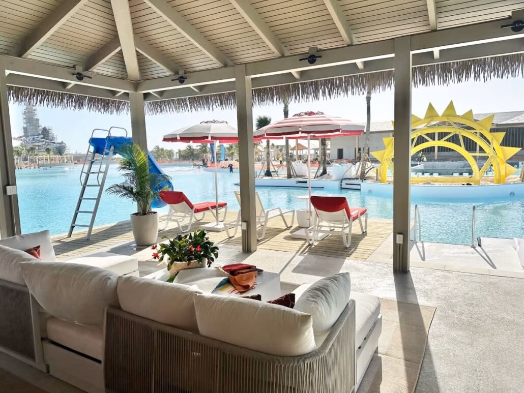A poolside view from under a shaded cabana with a sofa and flowers, capturing the vibrant essence of a Bahamas celebration. The area boasts lounge chairs with red cushions, umbrellas, a small water slide, and a large yellow sun-shaped structure in the water. Bright carnival-like weather prevails.