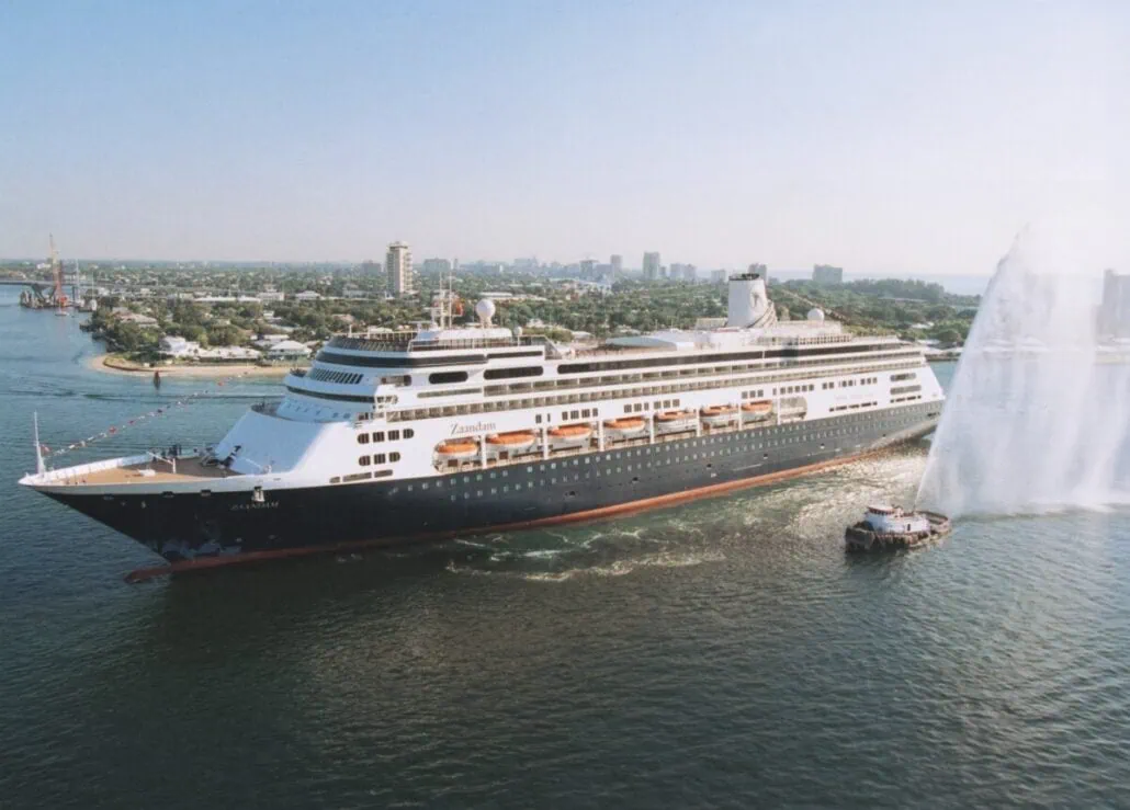 A large cruise ship sails near the shore, accompanied by a smaller boat spraying water. The background shows a city skyline with buildings and green trees under a clear blue sky.