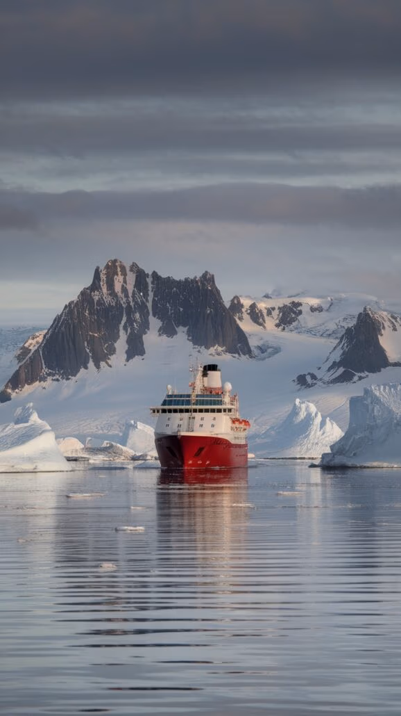 A red ship explores icy waters on a Northern Europe cruise, with snow-covered mountains towering in the background. The overcast sky casts a serene light, while the calm sea mirrors the landscape's beauty. Icebergs drift around this incredible destination.