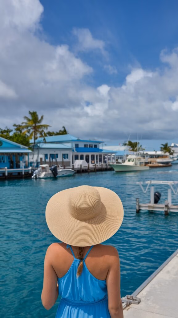 A person wearing a wide-brimmed straw hat and a blue dress stands on a dock, overlooking a turquoise marina, perhaps checking their cruise packing list. In the background, boats and blue-roofed buildings bask under the partly cloudy sky.