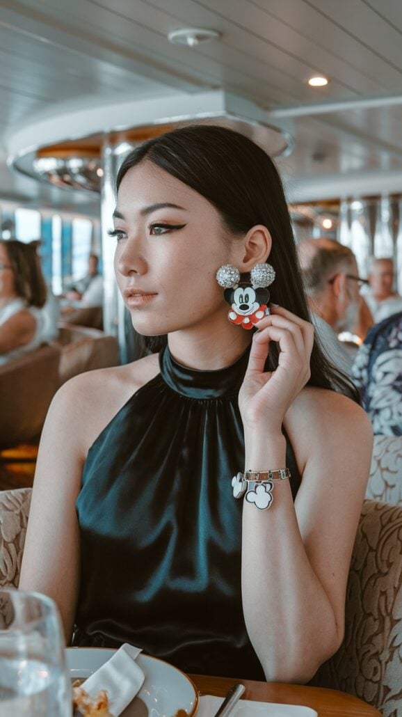 A woman in a sleeveless black halter dress holds up a Mickey Mouse earbud case with her right hand. Her Disney Cruise outfit is complete with matching Mickey Mouse-themed earrings and bracelet, all perfect magical picks. She sits indoors, surrounded by others in the background.