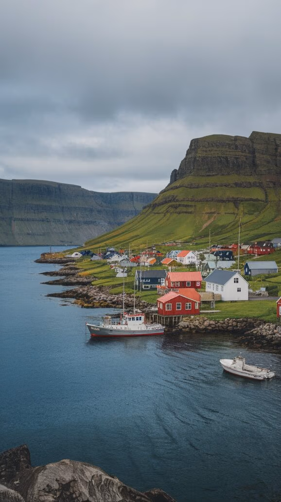 Nestled along a rocky shoreline, this scenic coastal village in Northern Europe is an incredible destination to explore. Its colorful houses, including a prominent red one, overlook two boats docked in calm waters, while hills with green and brown grass rise under a cloudy sky.