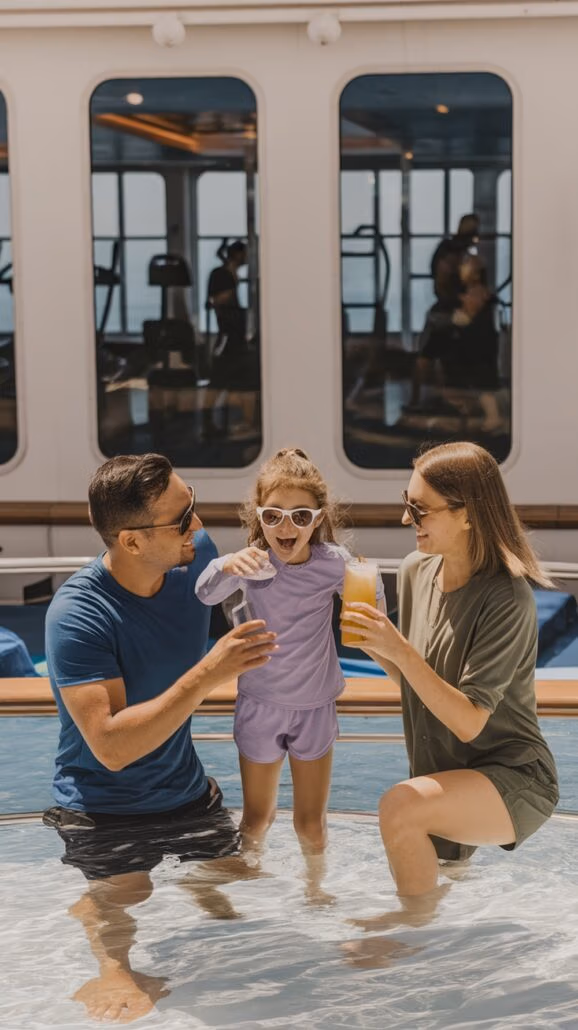 A man and woman kneel in a shallow pool, each holding a drink. Their child, in purple sunglasses, stands smiling between them. The large windows behind reveal gym equipment, hinting at the family's active lifestyle—one of the many reasons family-friendly Caribbean cruises are so popular with moms.