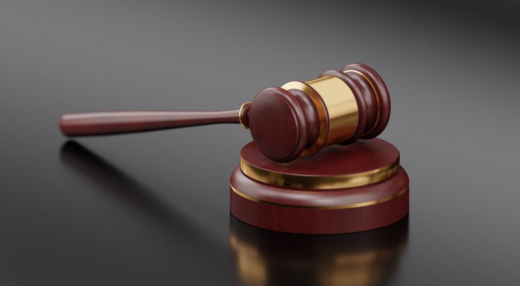 A wooden gavel with a gold band rests on a matching circular sound block on a dark surface, often symbolizing justice for swindling acts. The handle is smooth and polished, and the head is slightly larger on one end. The gavel is positioned diagonally across the sound block.